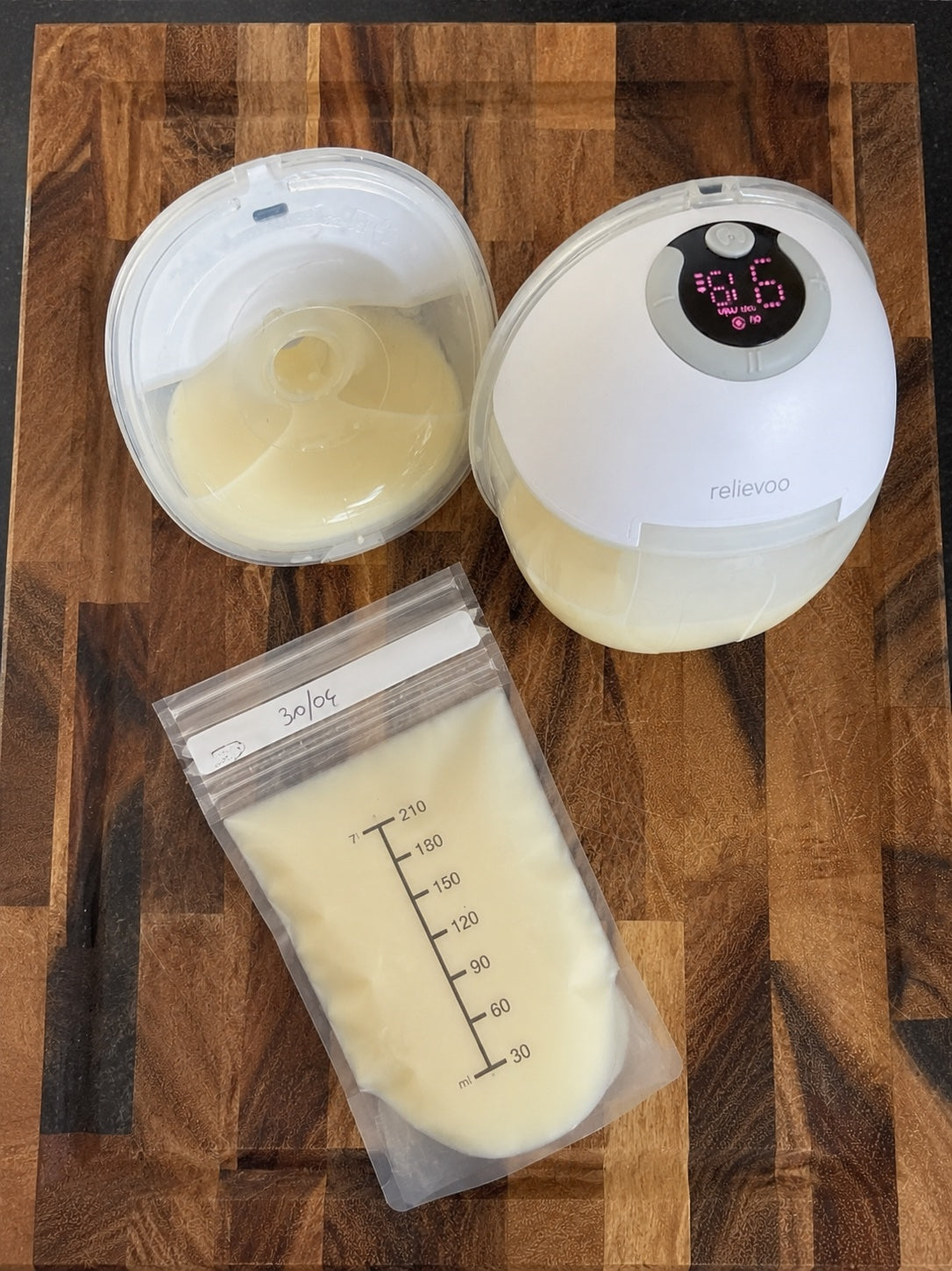 Relievoo wearable breast pump cup with fresh expressed breast milk next to a measurement-marked milk storage bag on a wooden kitchen board